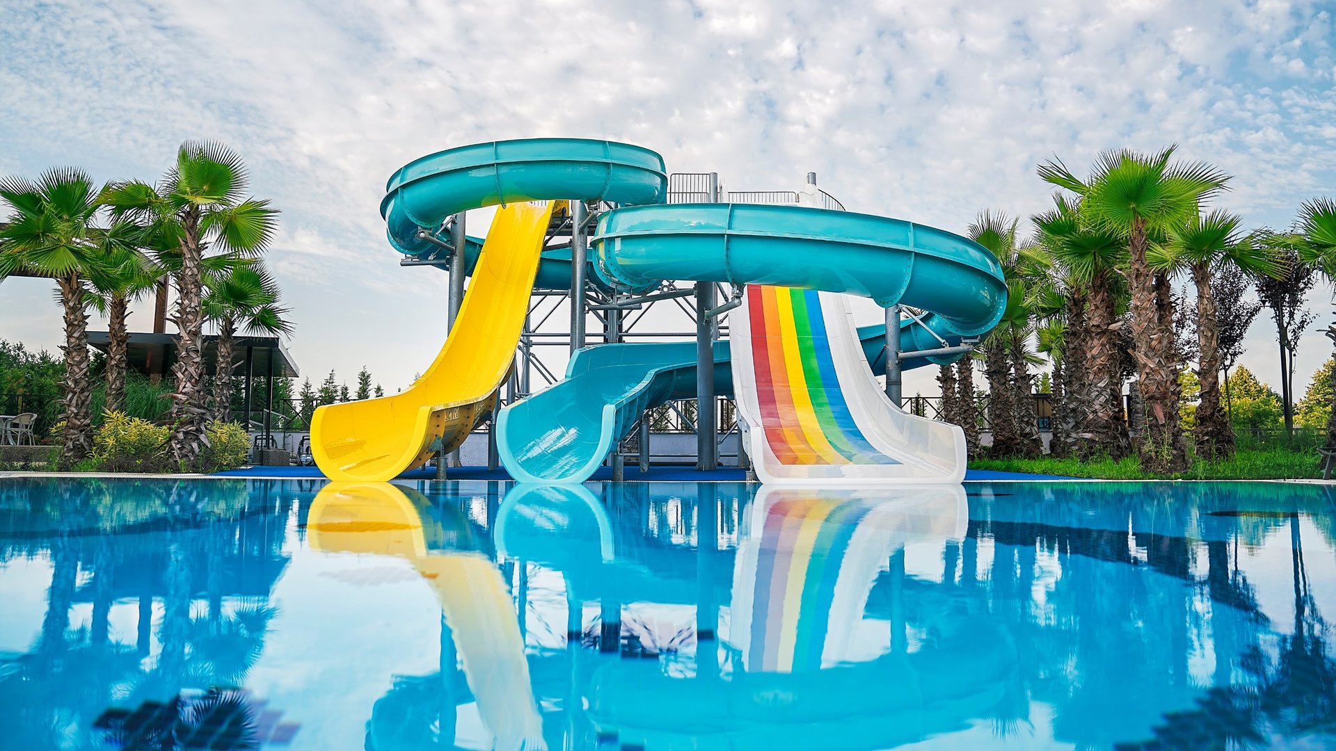 Colorful water slides at a tropical resort with blue pools, yellow and rainbow slides, and palm trees under a clear sky