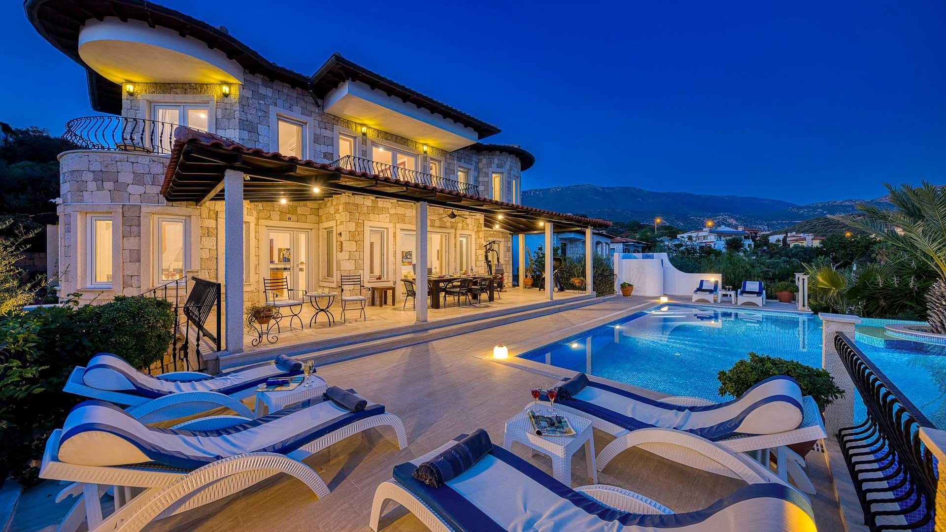 Modern luxury villa at dusk with illuminated house, infinity pool, and lounge chairs overlooking coastal hills